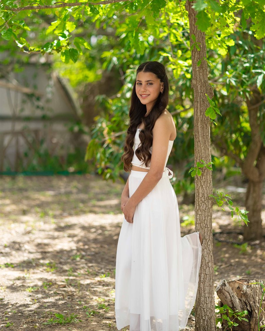 Young girl in a white dress smiling under green trees.